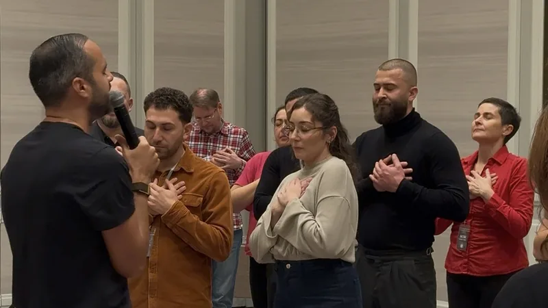 Darko Krstic spricht ins Mikrofon, die Teilnehmer stehen mit geschlossenen Augen und einer Hand auf der Brust